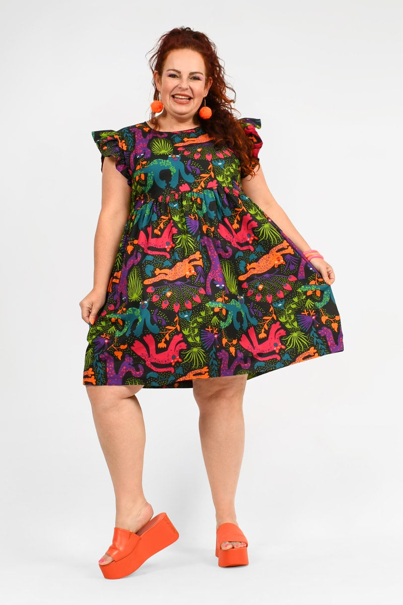 A woman modeling a colorful sundress with a animal print, including a dinosaur design, and cap sleeves. She is wearing orange open-toe platform shoes and has accessorized with red earrings.